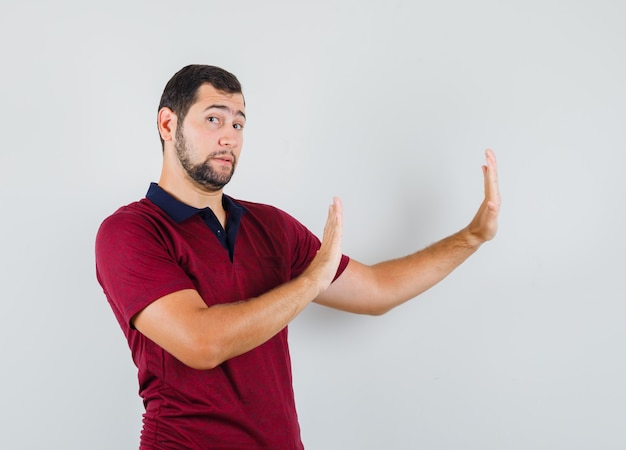 Young man showing stop gesture to aside in red t-shirt and looking troubled. front view.