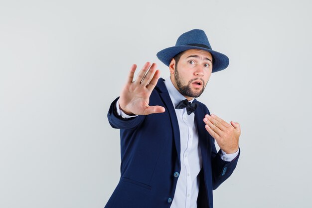 Young man showing stop gesture by pointing at himself in suit, hat and looking scared , front view.