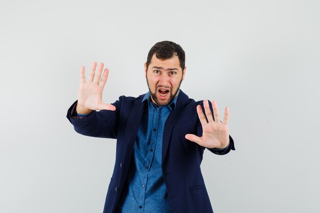 Young man showing stop gesture while screaming in shirt, jacket and looking frightened