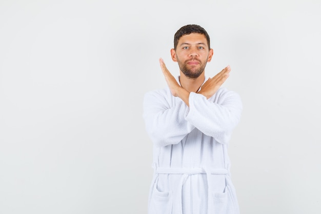 Young man showing stop gesture in white bathrobe and looking confident. front view.