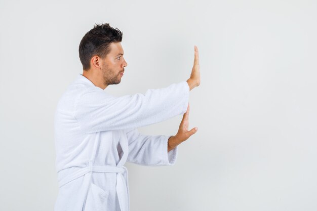 Young man showing stop gesture in white bathrobe and looking tranquil.