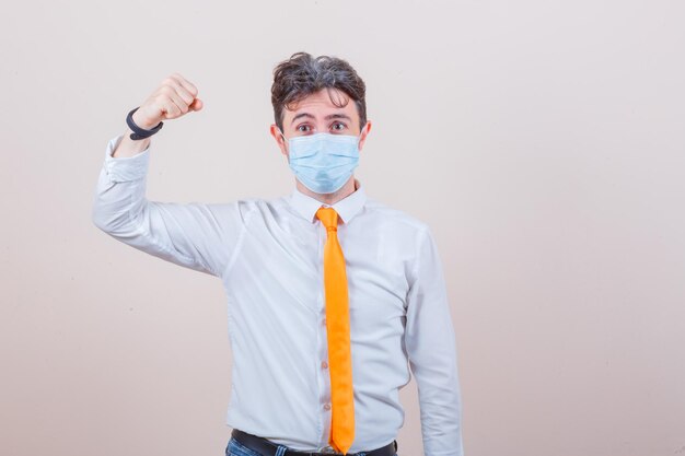 Young man showing success gesture in shirt, tie, jeans, mask