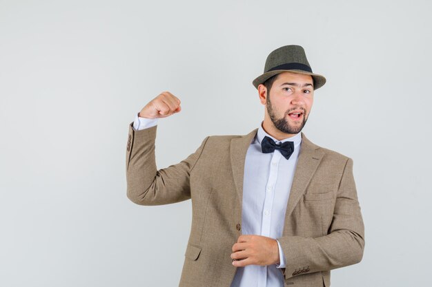 Young man showing success gesture in suit, hat and looking confident , front view.