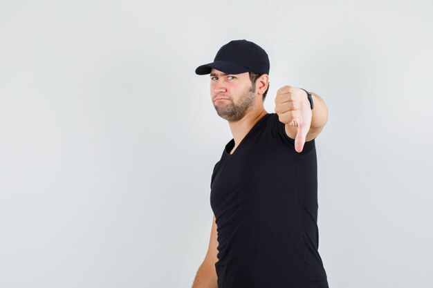 Молодой человек показывает палец вниз в черной футболке, кепке и выглядит разочарованным