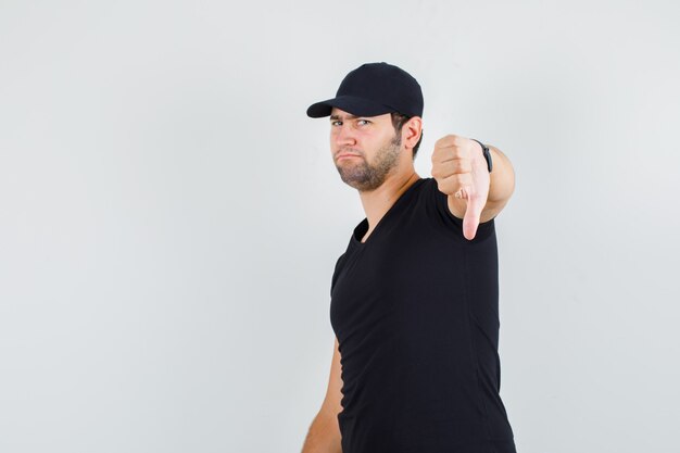 Young man showing thumb down in black t-shirt, cap and looking disappointed