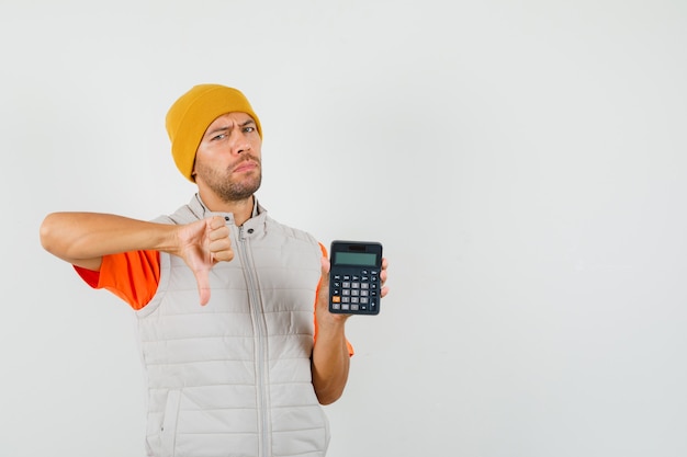 Young man showing thumb down while holding calculator in t-shirt, jacket, hat and looking dissatisfied.