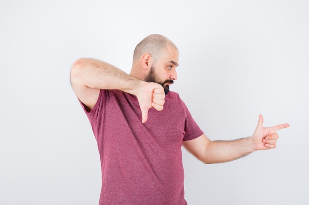 Young man showing thumb down while pointing aside in pink t-shirt front view.