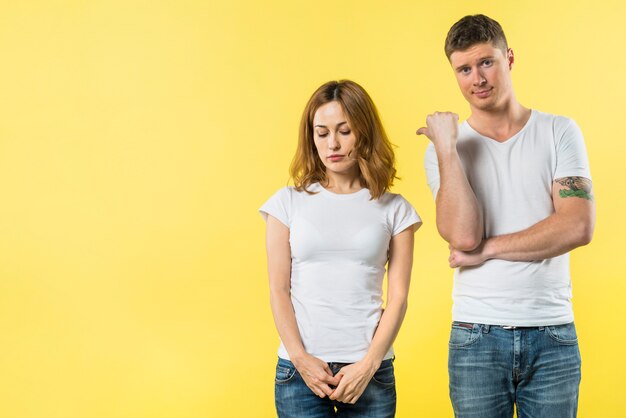 Young man showing thumb up to her sad girlfriend standing against yellow background