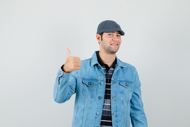 Young man showing thumb up in jacket,cap and looking glad , front view.