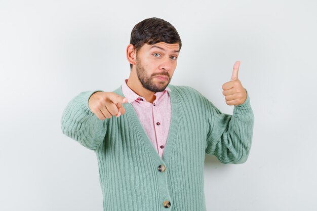 Young man showing thumb up, pointing forward in shirt, cardigan and looking pleased. front view.