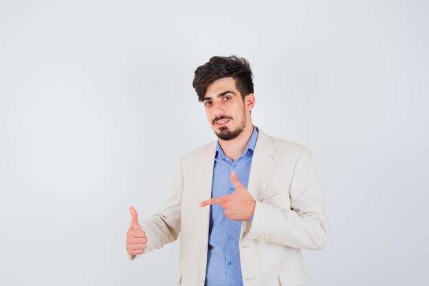 Young man showing thumb up and pointing to it in blue t-shirt and white suit jacket and looking serious