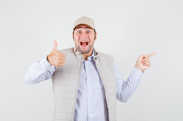 Young man showing thumb up and pointing right with index finger in beige jacket and cap and looking happy , front view.