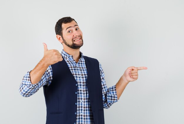 Young man showing thumb up, pointing to the side in shirt, vest and looking merry.