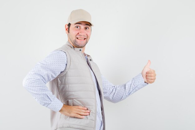 Young man showing thumb up and putting hand on waist in beige jacket and cap and looking happy. front view.