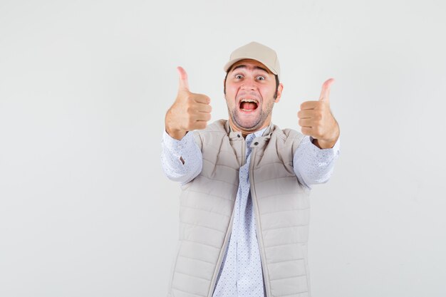 Young man showing thumb up in shirt,sleeveless jacket,cap and looking amused. front view.