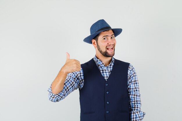 Young man showing thumb up in shirt, vest, hat and looking jolly.