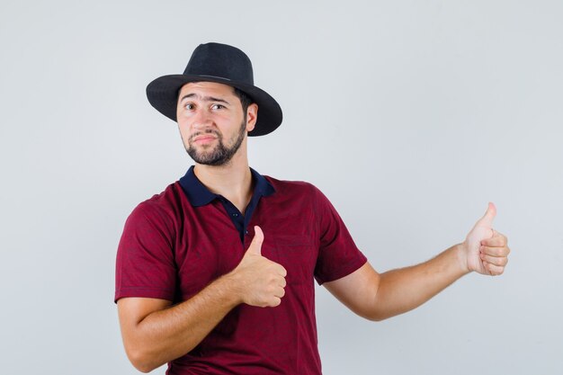 Young man showing thumb up at side in red t-shirt,hat and looking confident , front view.