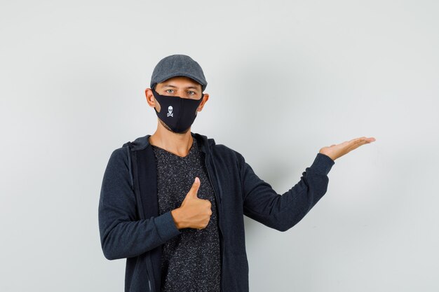 Young man showing thumb up, spreading palm aside in t-shirt, jacket, cap, mask