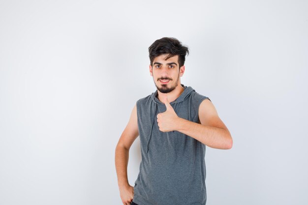 Young man showing thumb up while holding hand on waist in gray t-shirt and looking serious