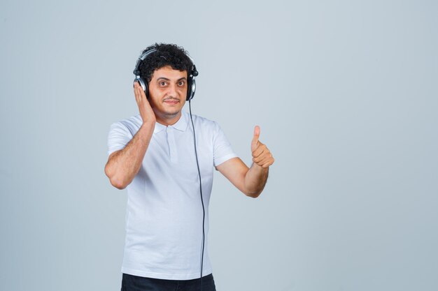 Young man showing thumb up while listening to music in white t-shirt and looking confident. front view.