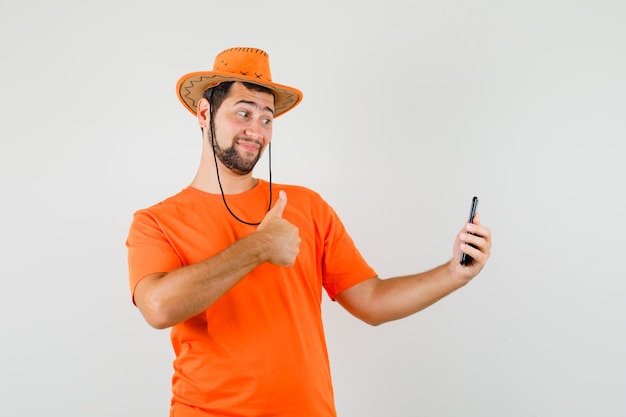 Young man showing thumb up while taking selfie in orange t-shirt, hat and looking happy. front view.