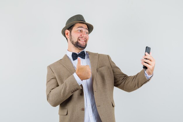 Young man showing thumb up while taking selfie in suit, hat and looking cheery , front view.