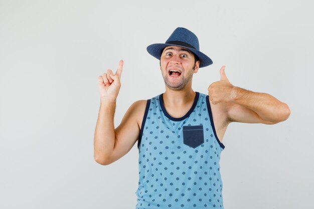 Молодой человек показывает палец вверх с пальцем в синей майке, шляпе и выглядит веселым. передний план.