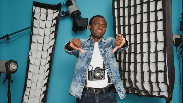 Free photo young man showing thumbs down dislike gesture in studio with professional photography equipment. expressing disagreement and disapproval, having tripod, and lighting backstage.