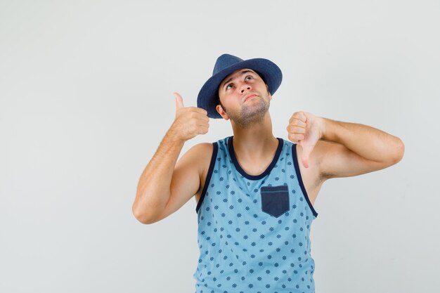 Young man showing thumbs up and down in blue singlet, hat and looking pensive. front view.