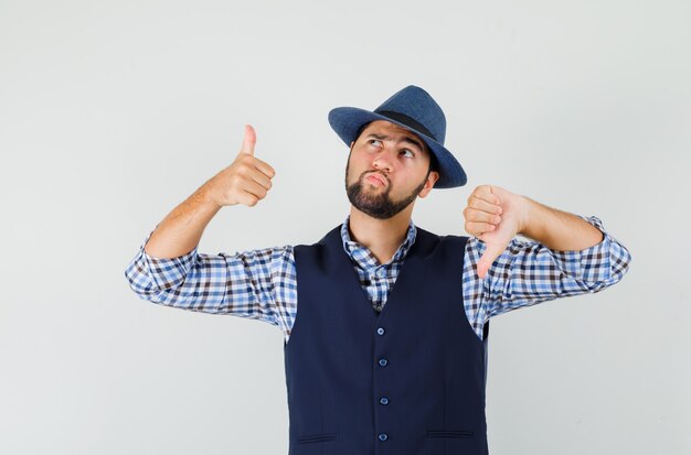 Young man showing thumbs up and down in shirt, vest, hat and looking indecisive.