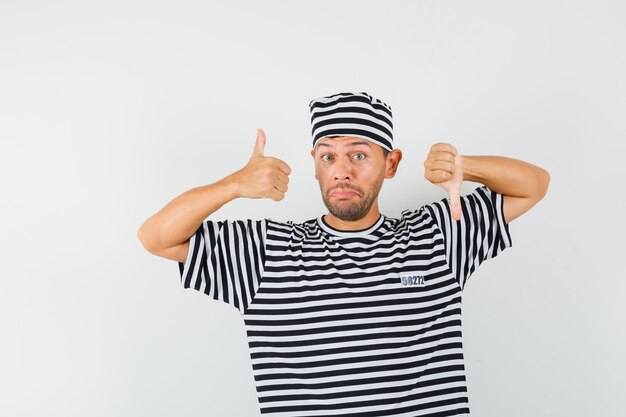 Young man showing thumbs up and down in striped t-shirt, hat and looking hesitant.