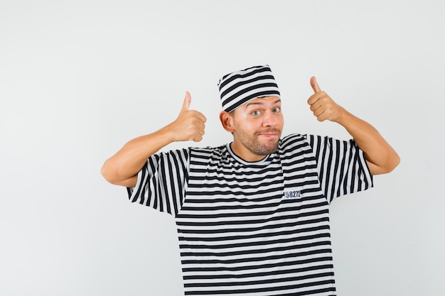 Young man showing thumbs up in striped t-shirt, hat and looking merry.