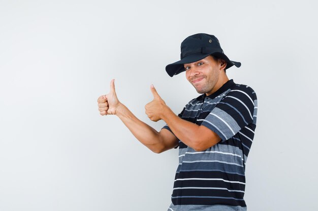 Молодой человек показывает палец вверх в футболке, шляпе и выглядит счастливым, вид спереди.
