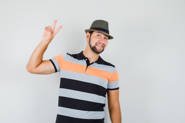 Молодой человек показывает v-знак в футболке, шляпе и выглядит веселым.