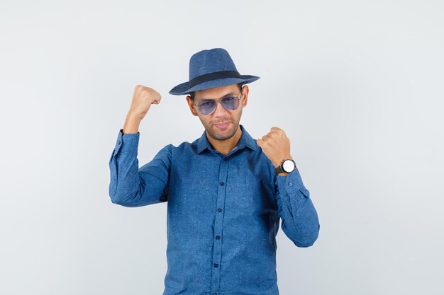Young man showing winner gesture in blue shirt, hat and looking happy. front view.