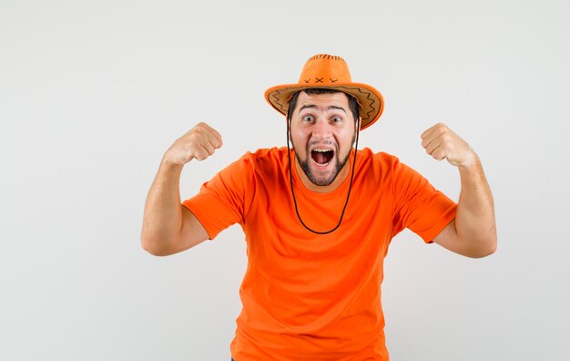 オレンジ色のTシャツ、帽子で勝者のジェスチャーを示し、至福に見える若い男。正面図。