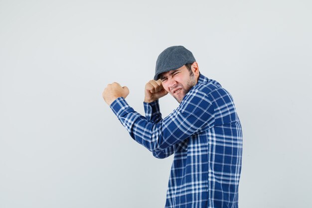 Young man showing winner gesture in shirt, cap and looking blissful .