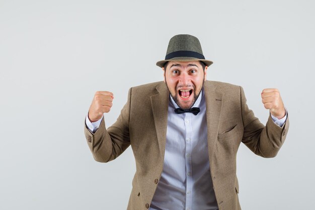 Young man showing winner gesture in suit, hat and looking blissful. front view.