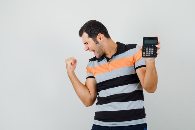 Young man showing winner gesture while holding calculator in t-shirt and looking cheerful. front view.