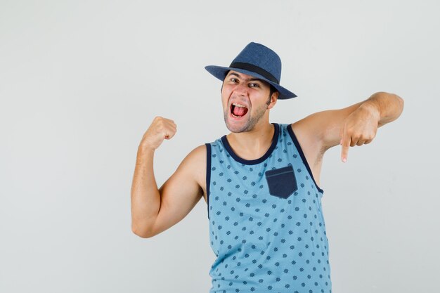 Young man showing winner gesture with finger down in blue singlet, hat and looking blissful , front view.