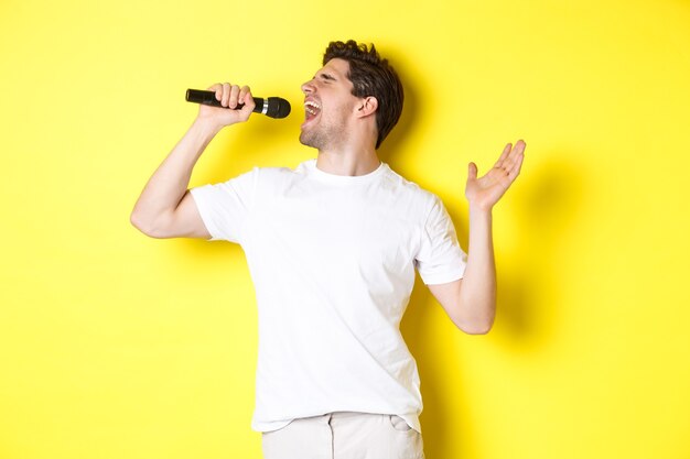 Певец молодой человек держит микрофон, достигая высокой ноты и поет караоке, стоя на желтом фоне.
