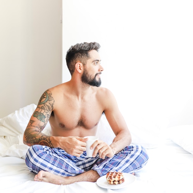 Young man sitting on bed with cup of coffee and sweet food on plate