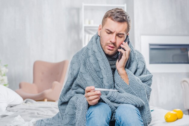 Young man sitting on bed wrapped in scarf looking at thermometer talking on mobile phone
