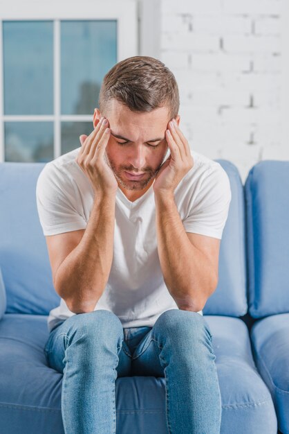 Молодой человек сидит на синем диване, чувствуя сильную головную боль, касаясь его лба