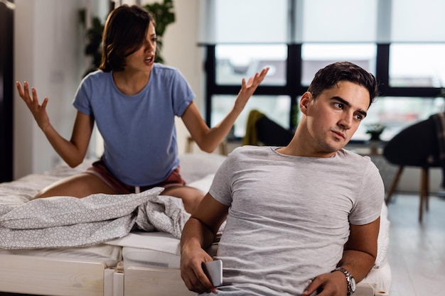 Free photo young man sitting by the bed and ignoring his girlfriend who is arguing with him