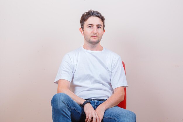 Молодой человек сидит на стуле и смотрит в камеру в футболке, джинсах