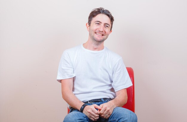 Young man sitting on chair in t-shirt, jeans and looking happy