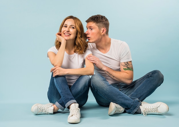 Free photo young man sitting on floor whispering in girlfriend's ear against blue background