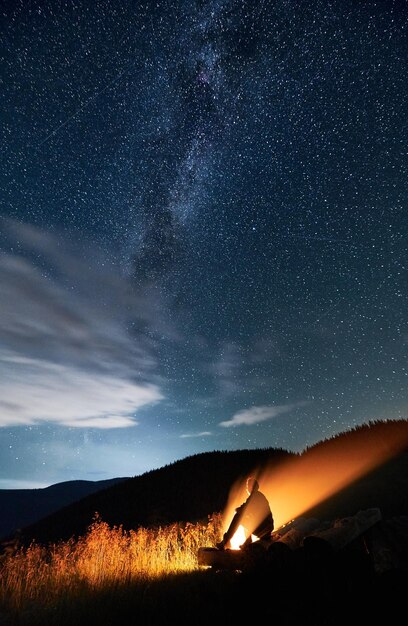 Young man sitting on logs near campfire in the mountains under sky full of stars