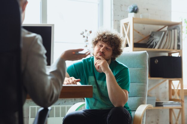 Young man sitting in office during the job interview with female employee, boss or HR-manager, talking, thinking, looks confident